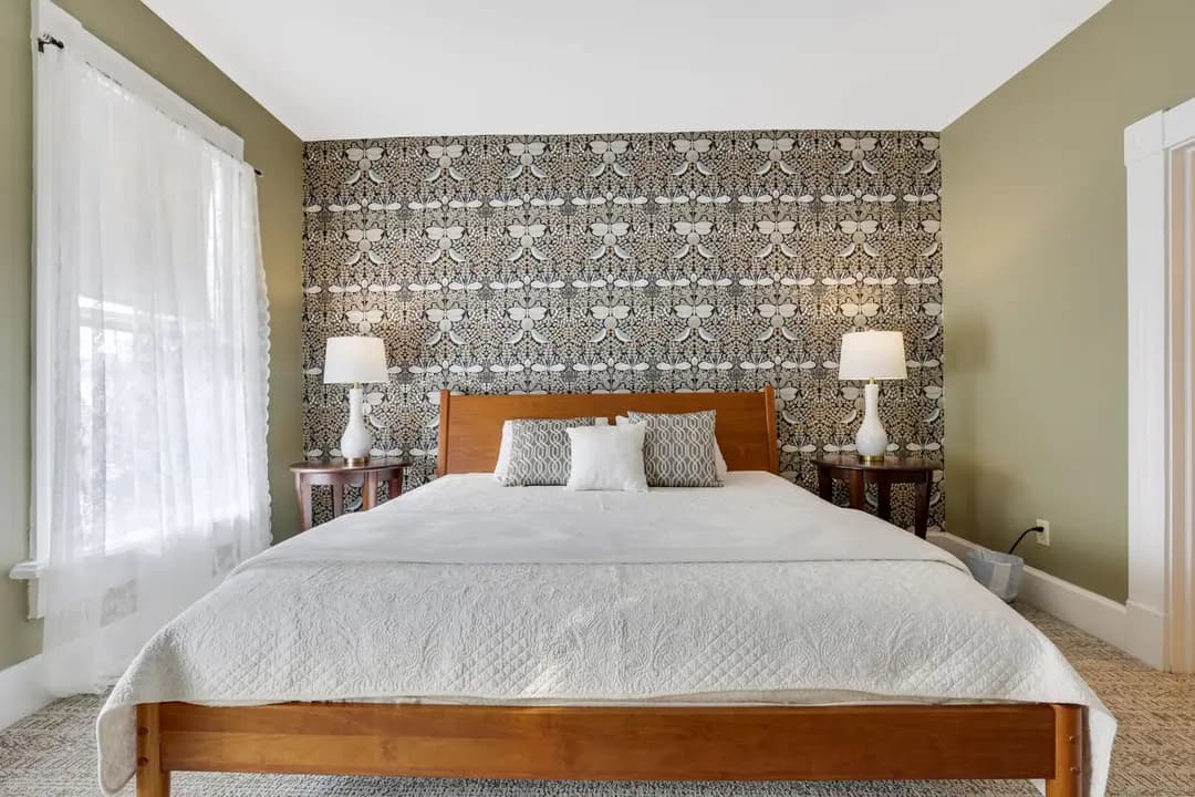 A cozy bedroom featuring a wooden bed with a white quilt, decorative pillows, and patterned wallpaper, illuminated by two bedside lamps.