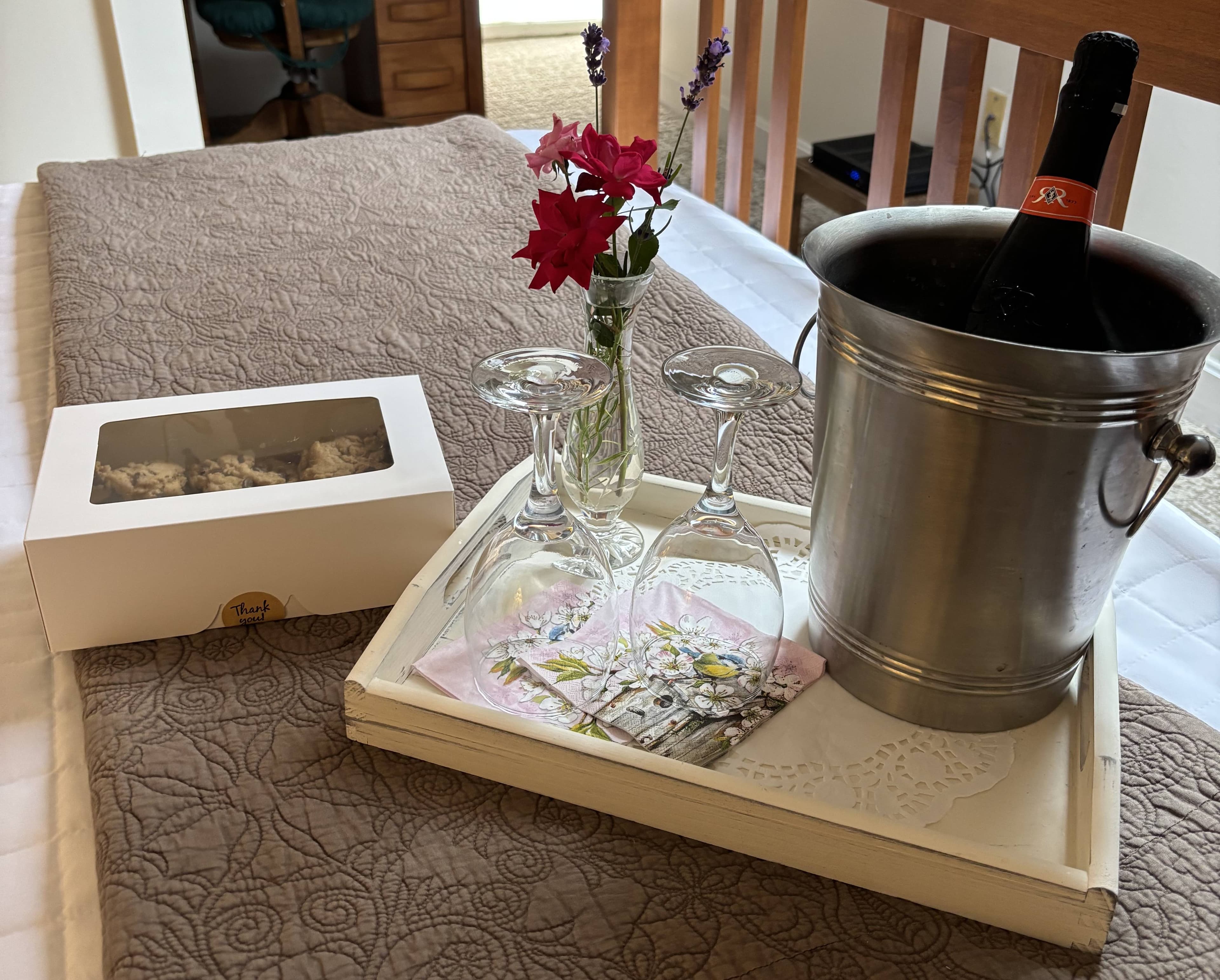 A tray with a chilled bottle of wine, two glasses, a small vase of flowers, and a box of treats rests on a bed with a quilted cover.