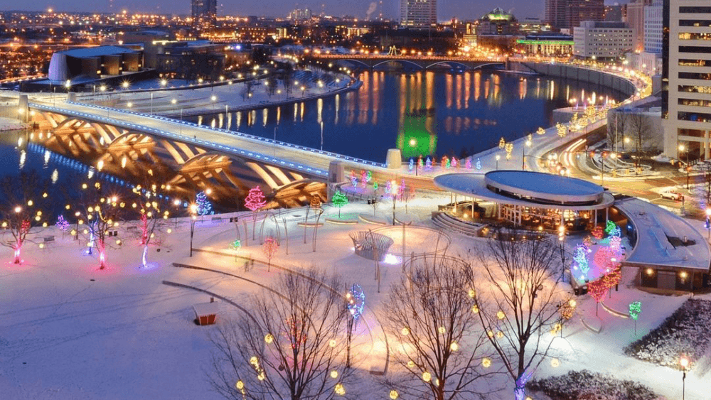A winter cityscape at night featuring a snow-covered park with colorful lights and a bridge over a shimmering river. A winter cityscape at night featuring a snow-covered park with colorful lights and a bridge over a shimmering river.