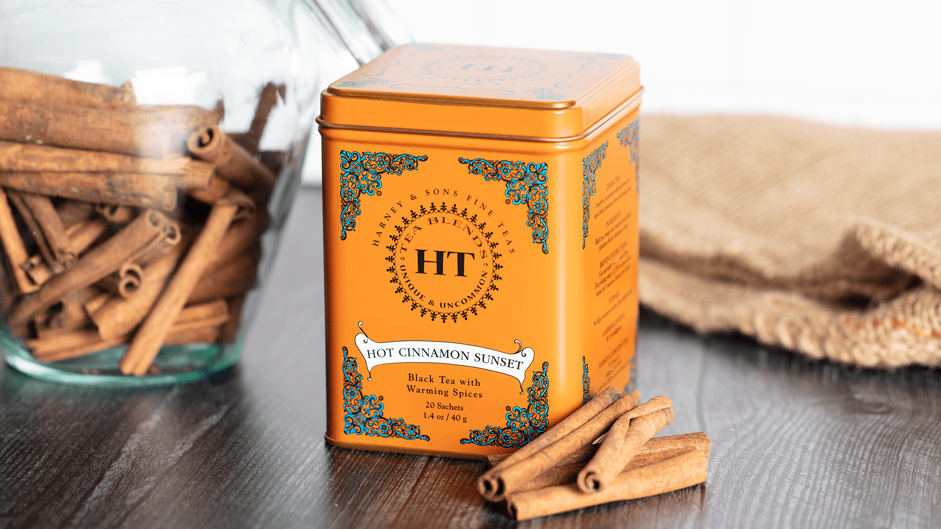 An orange tin of "Hot Cinnamon Sunset" black tea sits on a wooden surface, surrounded by cinnamon sticks and a glass jar filled with more sticks. An orange tin of "Hot Cinnamon Sunset" black tea sits on a wooden surface, surrounded by cinnamon sticks and a glass jar filled with more sticks.