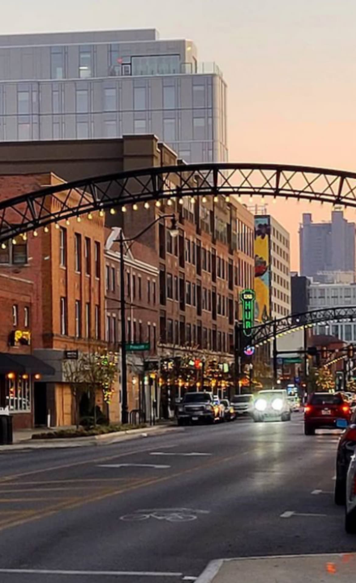 A lively urban street with historic buildings, warm lights, and a modern skyline at dusk.