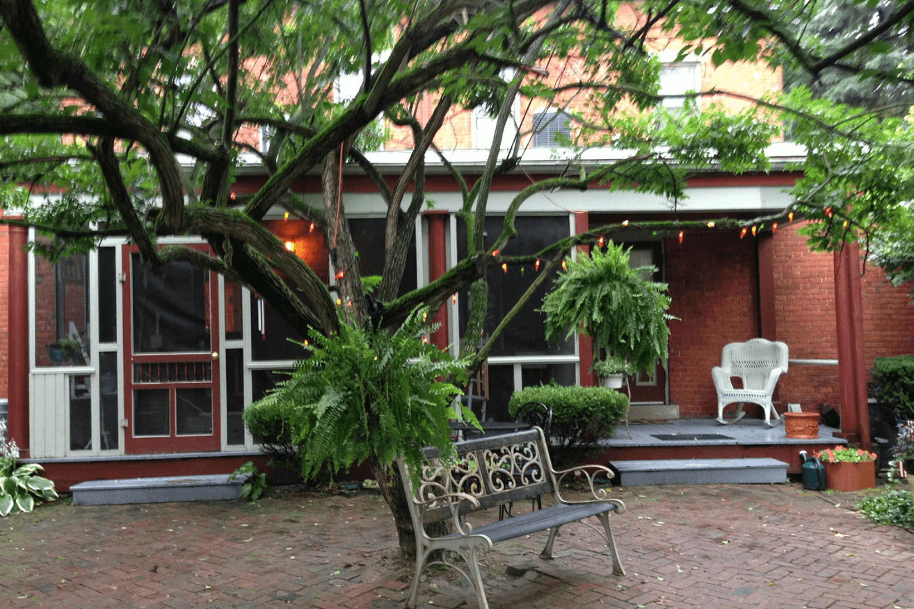 A brick patio backyard featuring a decorative bench in front of a tree in front of the back porch of a Victorian brick home.