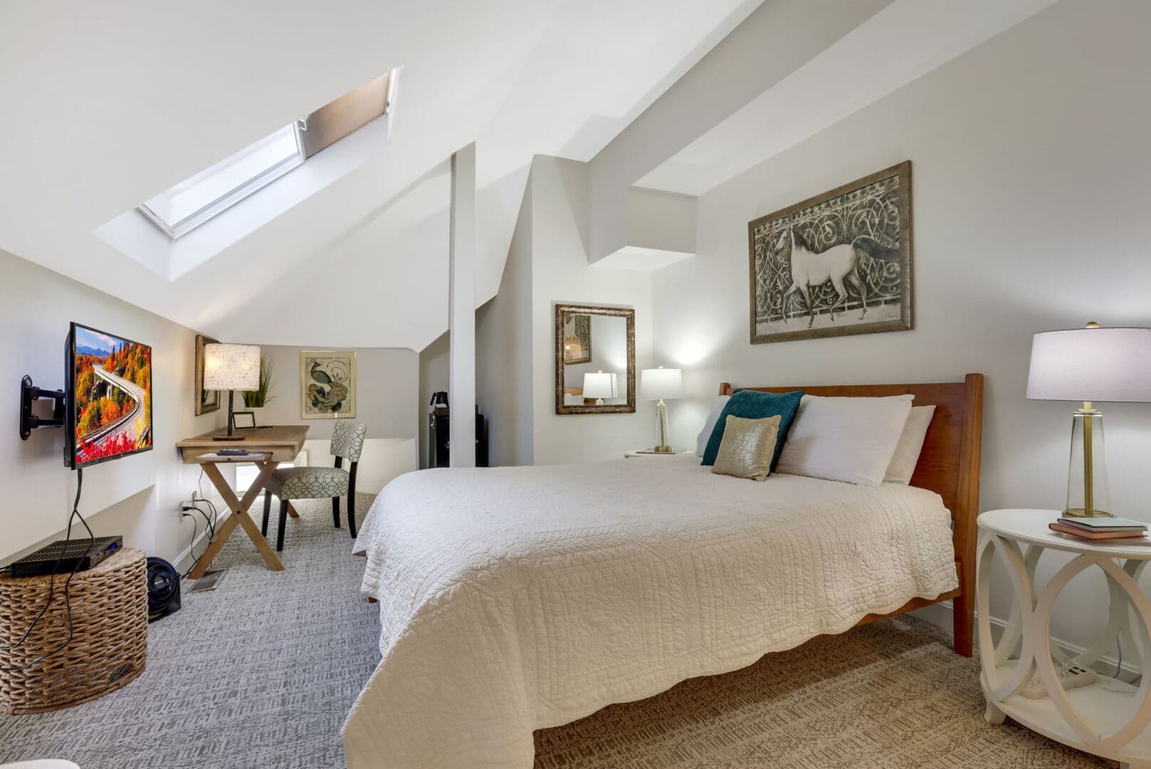A charming bedroom with a sloped ceiling and skylights, featuring a bed with white bedding, a TV, a desk, and decorative art.