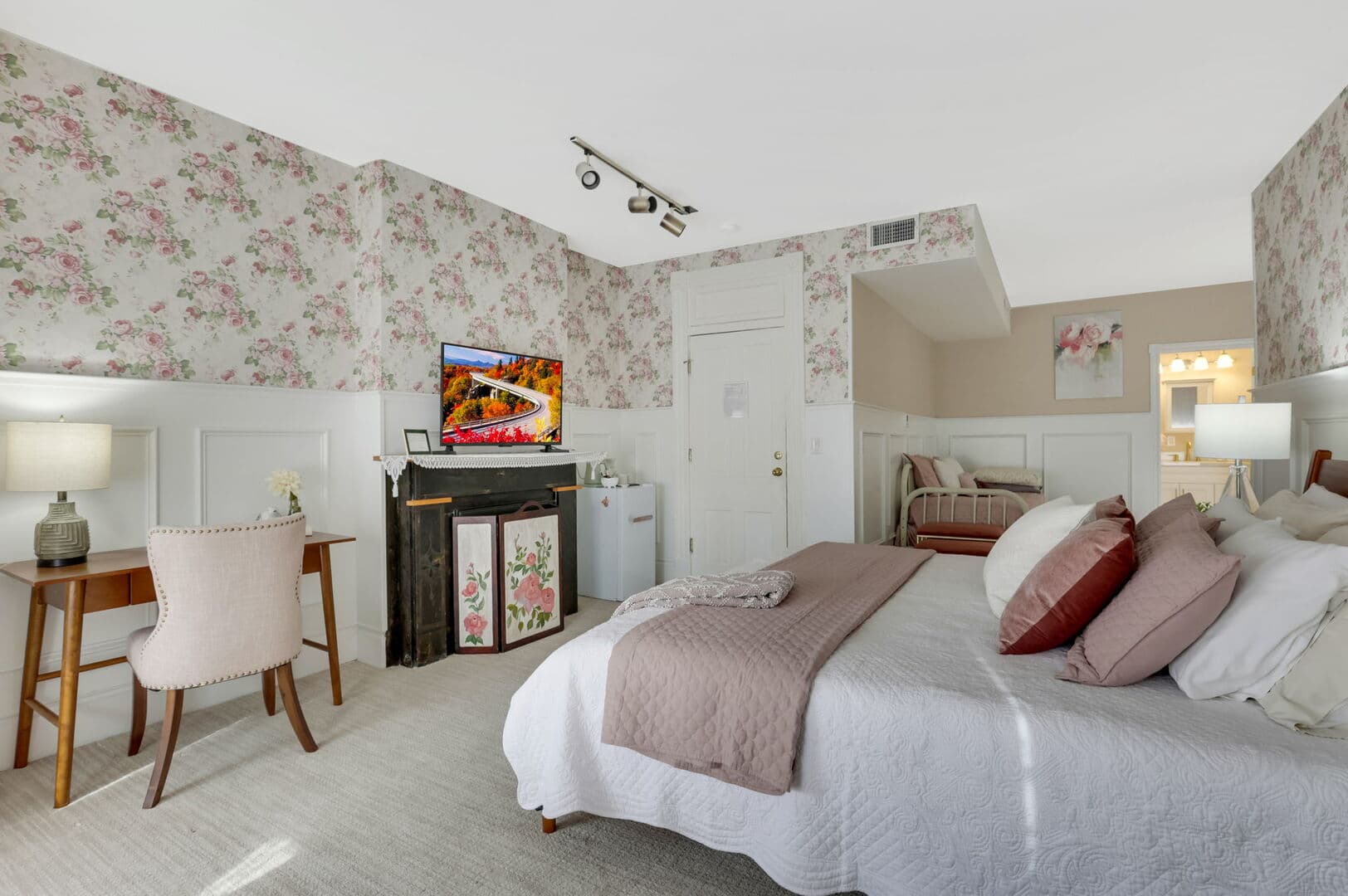 A charming bedroom with floral wallpaper and white wainscoting, featuring a queen bed with pink accents, a desk, a fireplace, and a TV.