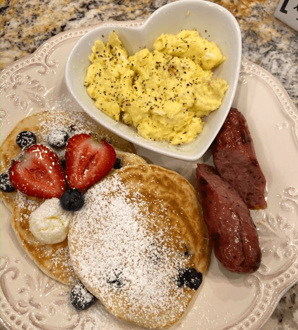 Breakfast entree featuring scrambled eggs in a heart-shaped ramekin atop a china plate with two strips of bacon and pancakes topped with powdered sugar, a pat of butter, sliced strawberries, and a few blueberries on a beige marble countertop