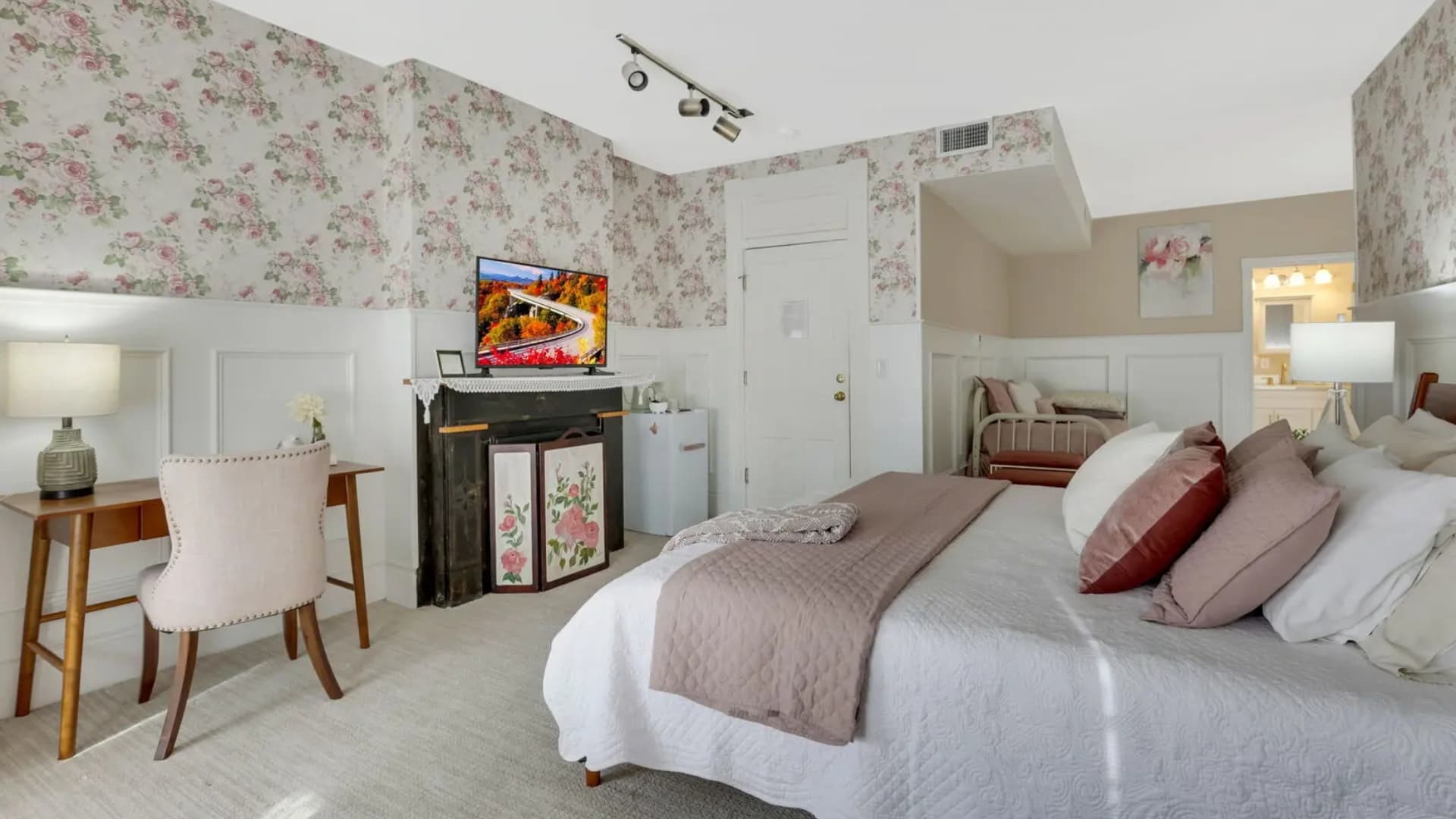 A cozy bedroom featuring floral wallpaper, a large bed with decorative pillows, a desk, a TV, and a small fridge.