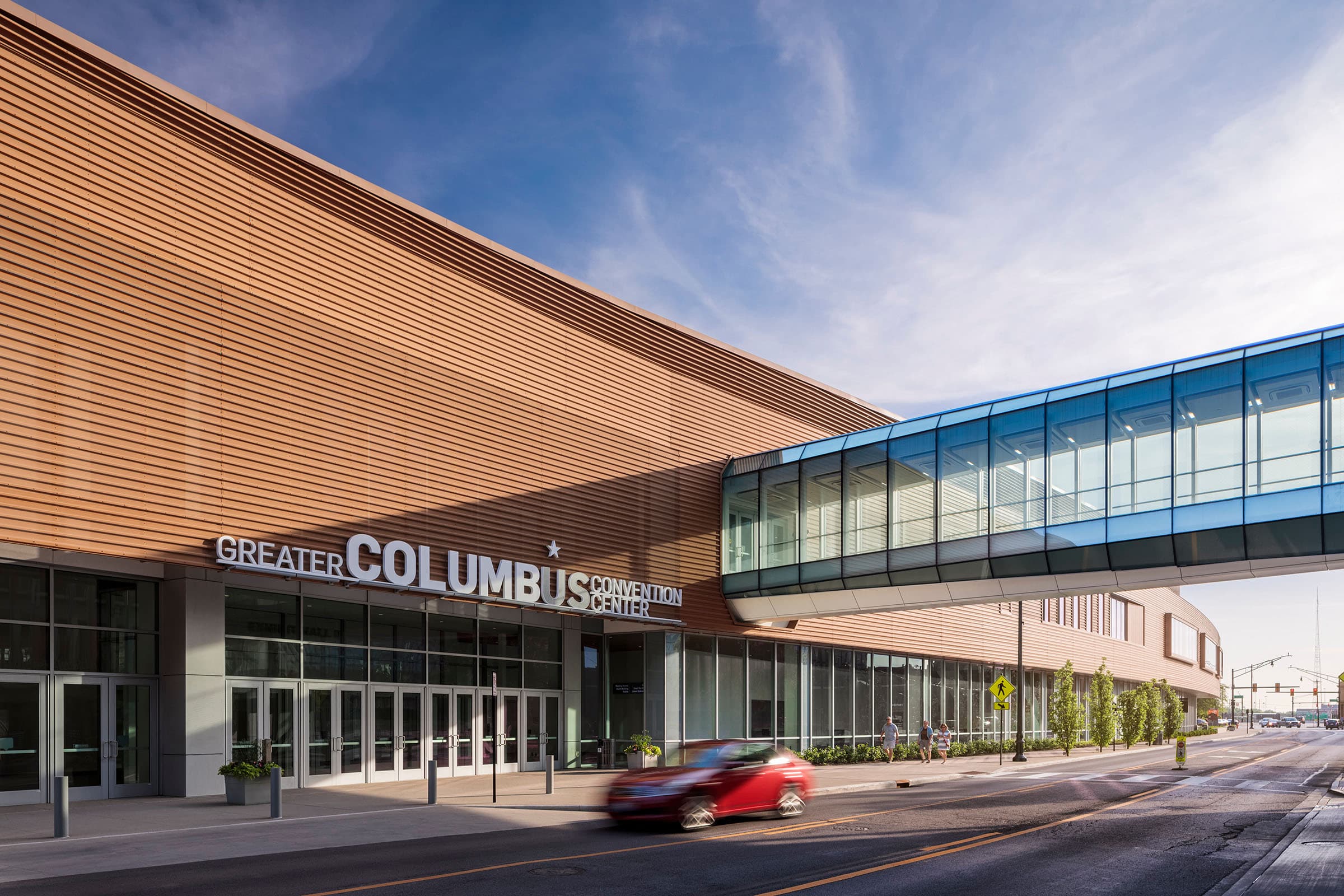 Exterior of the Greater Columbus Convention Center with a glass pedestrian bridge and a red car passing by.