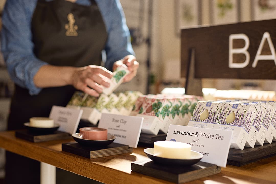A person arranges packaging for various soap products on a wooden display in a shop. A person arranges packaging for various soap products on a wooden display in a shop.