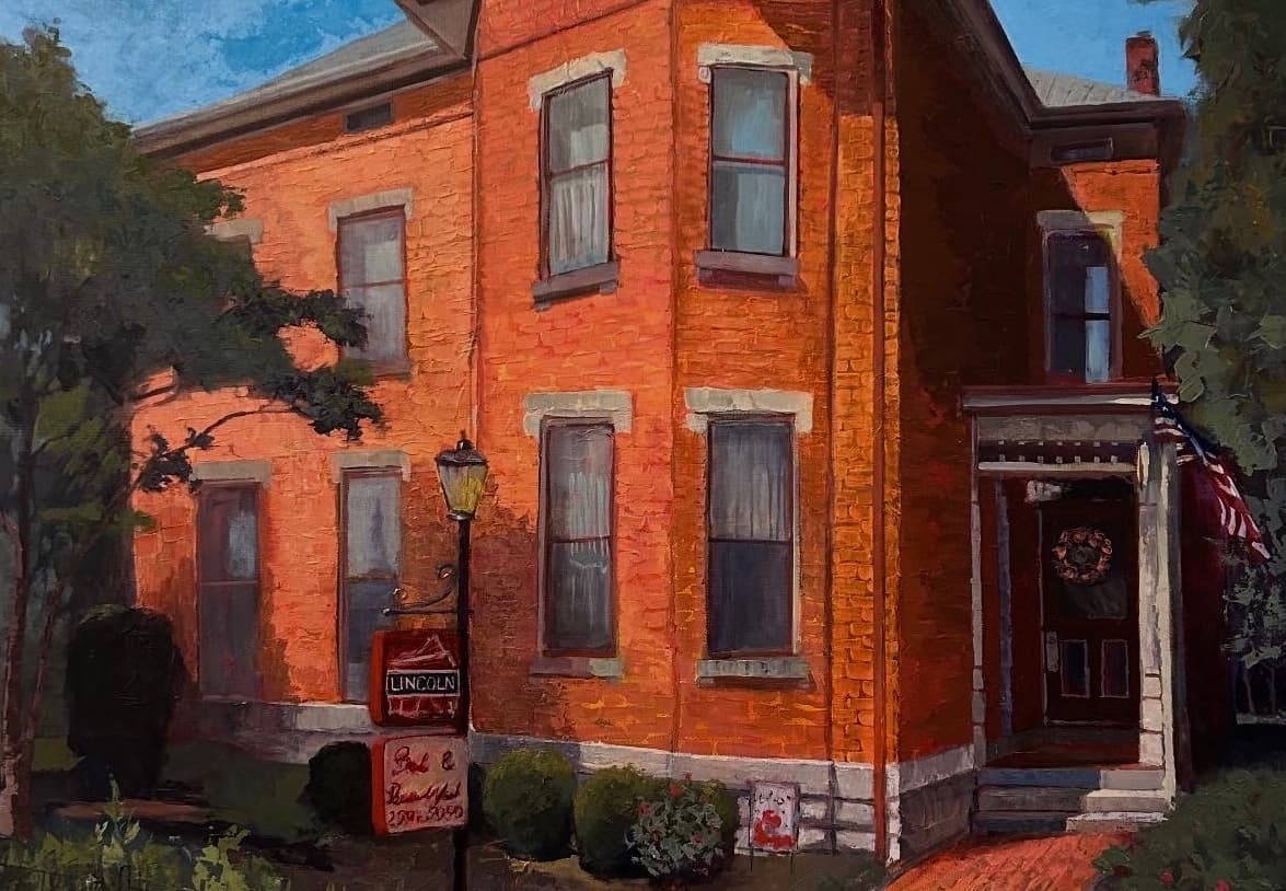 A historic brick house with white trim, featuring a welcoming entrance and a lamp post, surrounded by greenery.