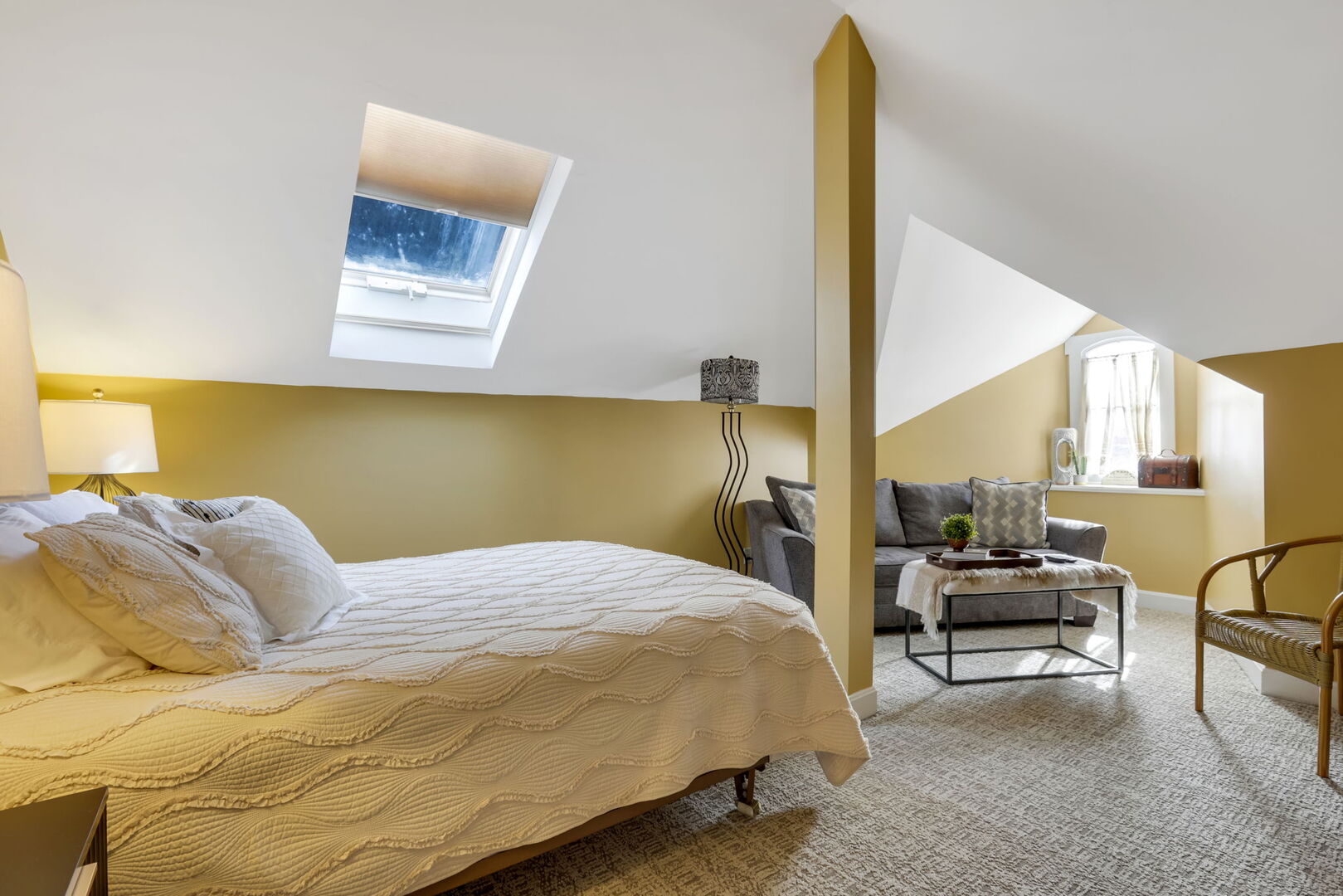 A sunlit third-floor bedroom with a skylight, featuring a queen bed, a cozy sitting area with a sofa and coffee table, and warm yellow accents.