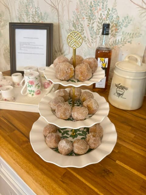 A tiered stand holds sugar-coated doughnut holes, with a framed document and a bottle in the background.