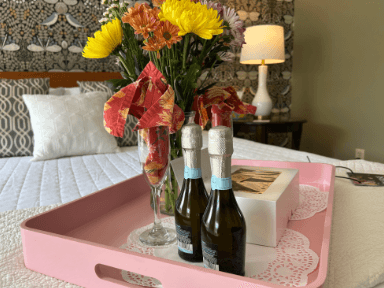 A pink tray holds two bottles of sparkling wine, a glass with flowers, and a gift box on a bed with a patterned wall in the background.